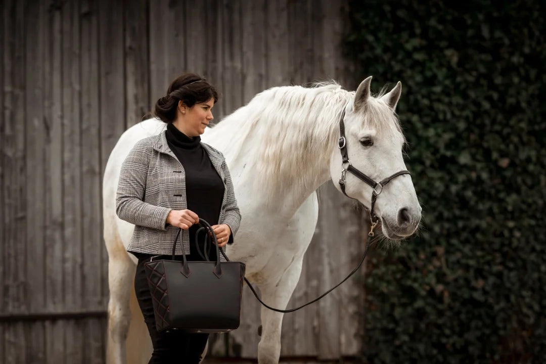 Elegant woman carrying a black leather hand bag by alpha pajé. She is accompanied by a white horse.