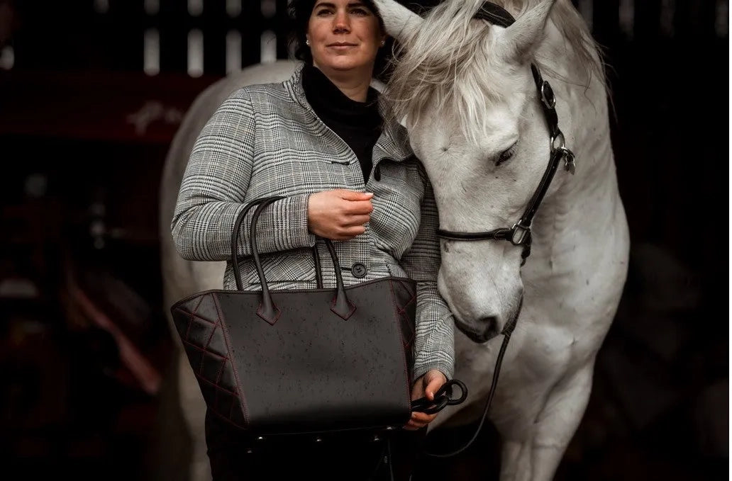 Elegant woman carrying a black luxury handbag by alpha pajé. She is accompanied by a white horse touching her arm.