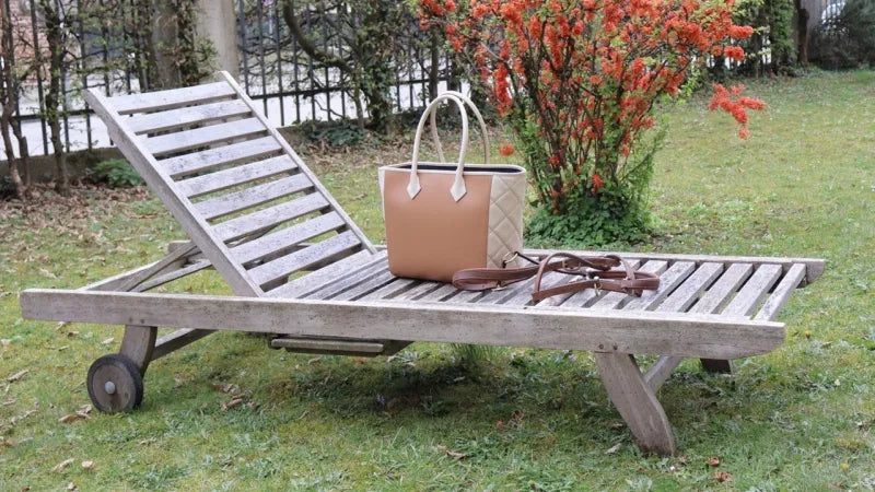 Brown luxury handbag on a garden lounger. In the background, a red flowering bush can be seen.